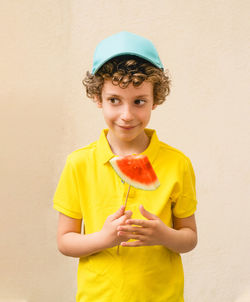 Smiling boy holding watermelon while standing against wall