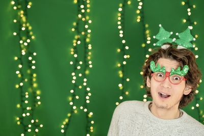 Surprised young man in new year's glasses on the background of a green wall with new year's lights. 