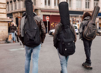 Rear view of friends carrying guitar case walking on street against buildings
