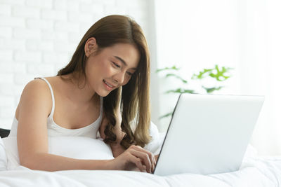 Smiling young woman using laptop while lying on bed at home