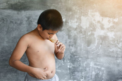 Shirtless boy eating popsicle while standing against wall