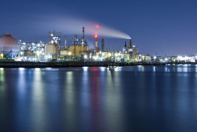 Illuminated factory by sea against sky at night