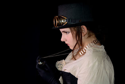 Young girl in steam punk look, from the side, looks at her watch, with black background