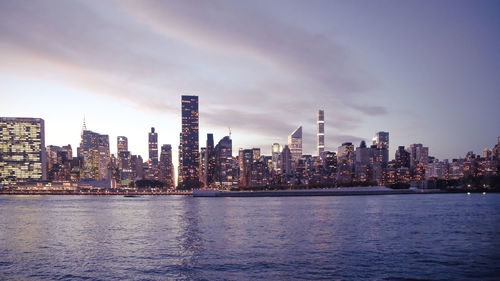 Modern buildings by river against sky in city during sunset