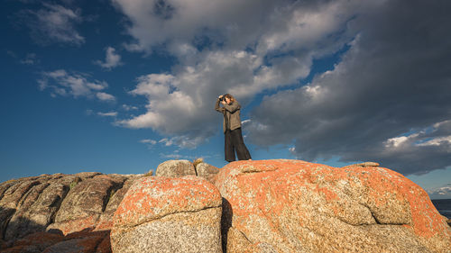 Low angle view of young man standing on rock against sky