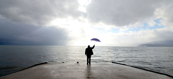 Rear view of man with umbrella standing by sea against cloudy sky