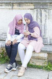 Young woman showing mobile phone to female friend while sitting on concrete bench