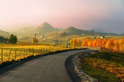 Country road leading towards mountains against sky during foggy sunrise