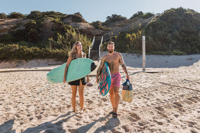 Full body of happy sportive couple with surfboards strolling together on sandy beach before training in the sea