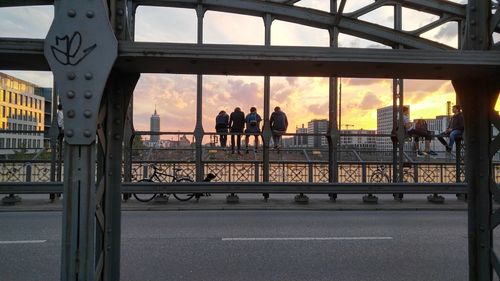 People sitting on bridge railing in city during sunset
