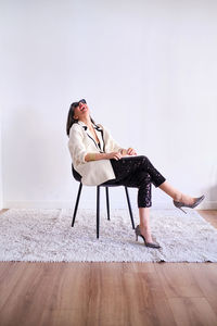 Full body of positive female in trendy outfit and high heels sitting on rug near white wall in light studio