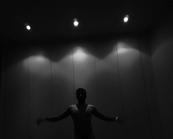 Rear view of shirtless man standing against illuminated wall