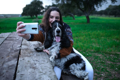 From above of happy young woman smiling during hug with black and white spaniel while shooting selfie on mobile phone in park