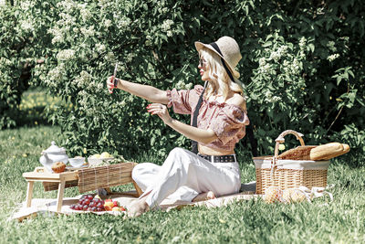 A young blonde woman in a hat and sunglasses at a picnic in the park .takes a selfie on a smartphone
