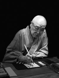 Senior man writing at table against black background