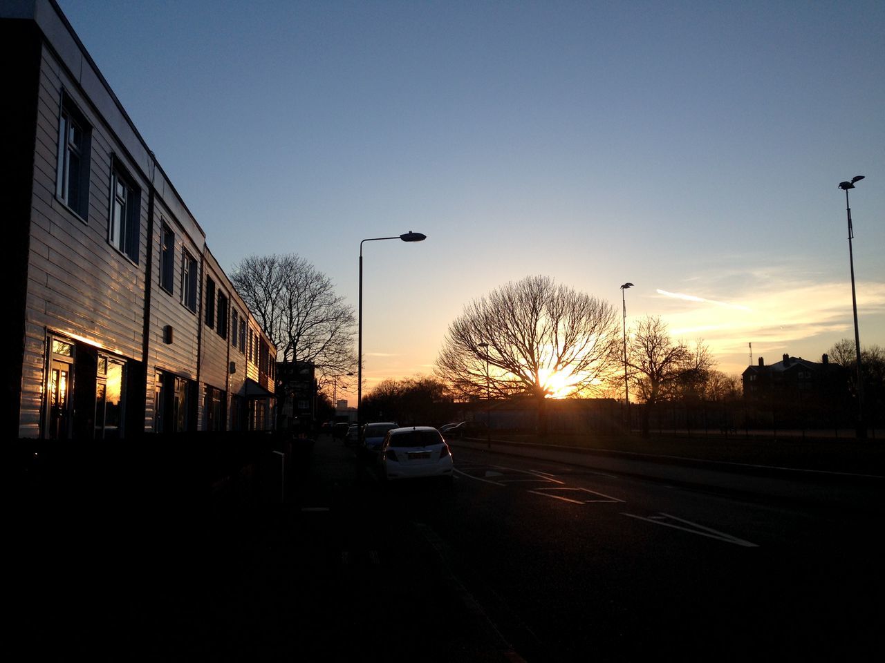 sunset, street light, transportation, building exterior, silhouette, car, street, built structure, sun, road, clear sky, architecture, sky, city, sunlight, the way forward, land vehicle, orange color, mode of transport, tree