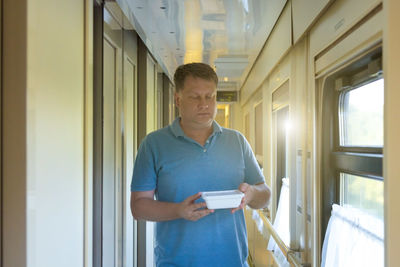 An adult blond man walks slowly along the train carriage carrying hot instant noodles.