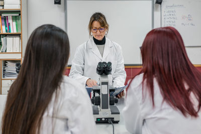 Adult female in white robe reading notes near microscope while teaching young women in modern laboratory in university