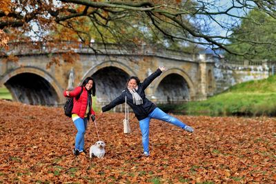 Women with arms outstretched standing with dog against bridge during autumn