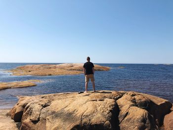 Rear view of man standing on rocky shore against clear blue sky