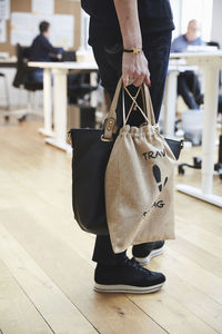 Low section of fashionable senior businesswoman standing with purse and bag on hardwood floor at office