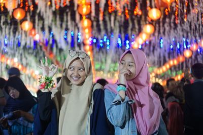 Smiling friends wearing hijab standing against illuminated lights at night