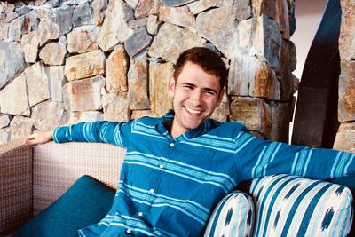 Smiling man looking away while sitting on sofa against stone wall