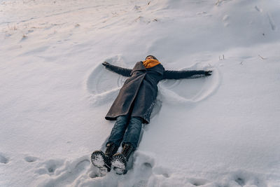 A woman in a coat, scarf and boots lies in a snow-covered field and makes a snow angel