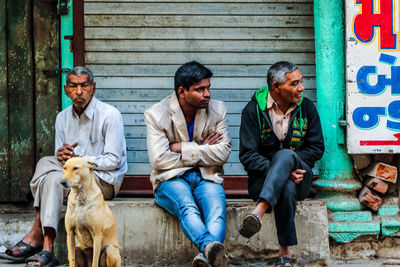 Men looking away while sitting on seat against closed shop