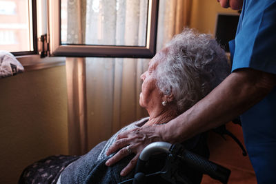 Side view of copped unrecognizable caring nurse in uniform standing near sick elderly female in wheelchair near window in light room