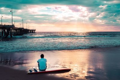 Rear view of man sitting on shore at sunset