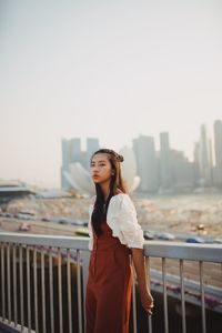 Portrait of young woman standing against railing with clear sky in background