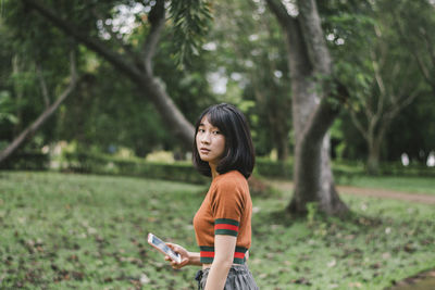 Portrait of young woman holding mobile phone while standing against trees in park