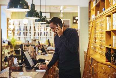 Confident young male owner talking on smart phone while writing at checkout counter in furniture store