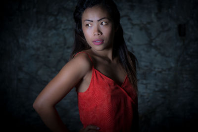 Young woman wearing red dress while looking away against wall in dark