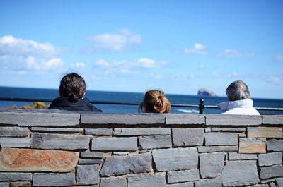 Rear view of people on retaining wall by sea against sky