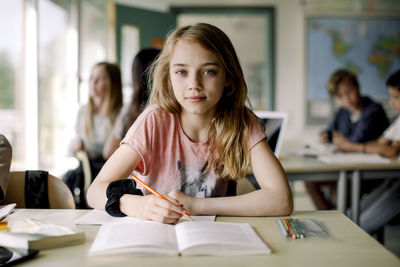 Portrait of female student writing in book while sitting at table in classroom