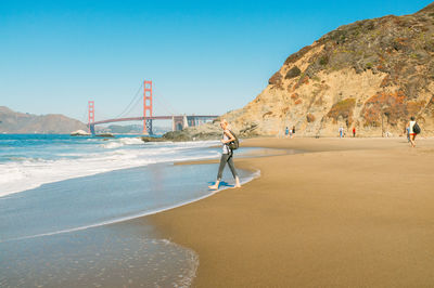 Woman walking on shore with golden gate bridge against clear sky