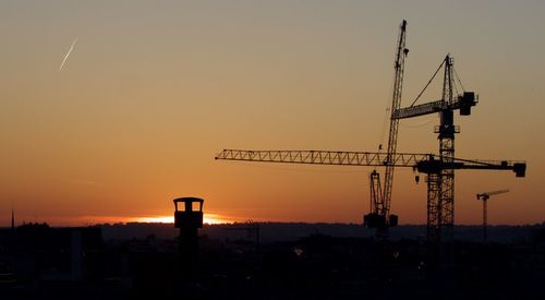 Silhouette cranes at construction site against sky during sunset