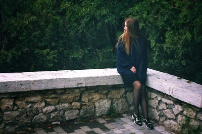 Full length of young woman sitting on retaining wall against trees