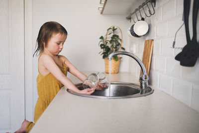 European child of 4 years old washes glass in sink on his own, bright kitchen in real interior, 