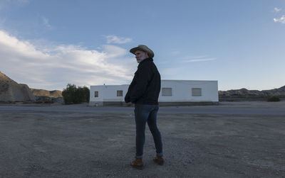 Adult man in cowboy hat standing in front of white building against sky