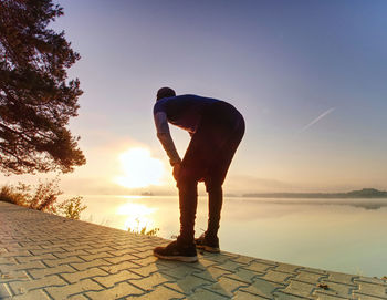 Short break for breath. silhouette of active man exercising and stretching on lake beach at sunrise