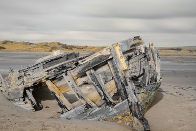 Damaged wooden boat at beach against cloudy sky