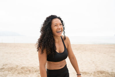 Positive asian female athlete with curly hair laughing on sandy seashore in summer