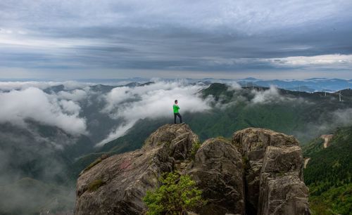 Man standing on cliff overlooking mountains and wind turbine