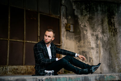 Young man looking away while sitting against old building