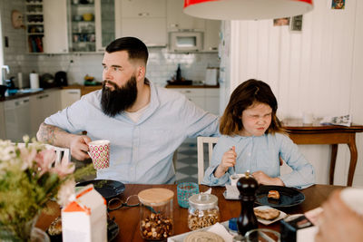 Father having drink while daughter making face during breakfast at home