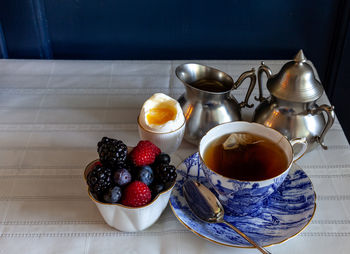 Tea, mixed berries and a soft boiled egg for breakfast on a table with a blue vase.