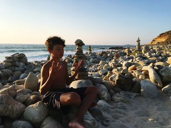 Shirtless boy with eyes closed meditating while sitting on rocks at beach against clear sky during sunset